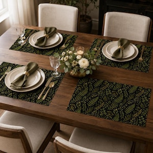Peut inclure: Une table à manger dressée pour quatre personnes avec des sets de table rectangulaires au motif de feuilles de fougère vert foncé et doré. Chaque couvert comprend une assiette en céramique blanche, une serviette en tissu vert olive avec un rond de serviette doré et des couverts dorés.