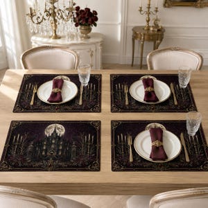 May include: A set of four rectangular placemats featuring a dark gothic castle illustration under a full moon, each topped with a white ceramic plate, a deep burgundy cloth napkin held by a gold filigree ring, a gold fork, a gold knife, and a glass.