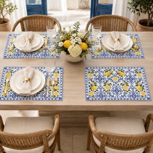 May include: A dining table set for four with rectangular placemats featuring a blue and white tile pattern with yellow lemons and green leaves. Each setting includes a white plate, a beige napkin with a gold ring, and gold-toned cutlery.
