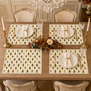 May include: A dining table set for four with cream-colored placemats featuring a repeating pattern of colorful autumn leaves and acorns. Each place setting includes a white ceramic plate, a folded white napkin with a gold ring, and silverware.