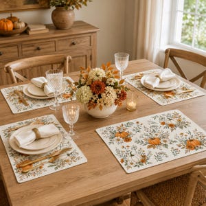 Könnte beinhalten: Ein Set rechteckiger Platzsets mit einem wiederkehrenden Muster aus orangefarbenen Kürbissen, weißen Blumen und grünem Blattwerk auf cremefarbenem Grund. Jedes Platzset ist mit einem weißen Teller, einer Serviette und goldenem Besteck gedeckt.