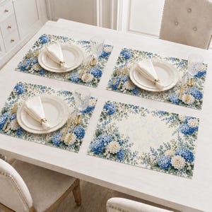 May include: Four rectangular placemats featuring a floral pattern of blue and white hydrangea blossoms and green leaves. Each setting includes a white ceramic plate, a folded white napkin with a gold ring, gold cutlery, and a clear glass.