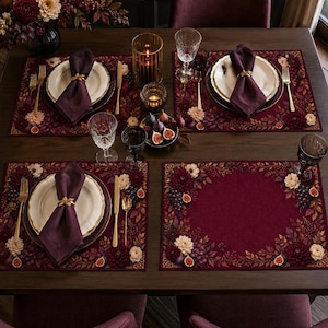 Peut inclure: Un ensemble de quatre sets de table rectangulaires bordeaux, ornés d'une bordure botanique de dahlias crème, de raisins noirs et de figues coupées. La table comprend des assiettes blanches à bordure dorée, des serviettes violettes et des couverts dorés.
