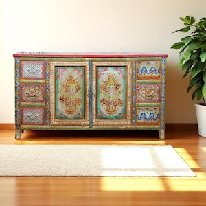 May include: A hand-painted wooden cabinet with floral designs in shades of green, orange, and pink. The cabinet has multiple drawers and doors, with a red top and silver legs. A white rug and a potted plant are in the foreground.