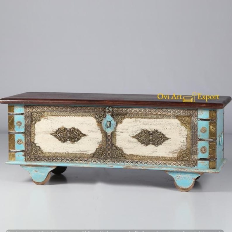 May include: A decorative wooden storage chest with a distressed white and blue paint finish. The chest has intricate metalwork details and brass accents. It features a hinged lid and sits on four small wheels.