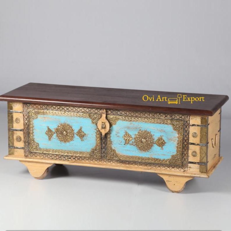 May include: A wooden storage chest with two doors, painted in a light cream color with blue accents and gold metal details. The top of the chest is made of dark wood.