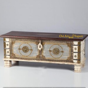 May include: A white wooden storage chest with gold accents and a dark wood top. The chest has two doors with ornate metal handles and a decorative design on the front. The text "Ovi Art Export" is visible on the top of the chest.