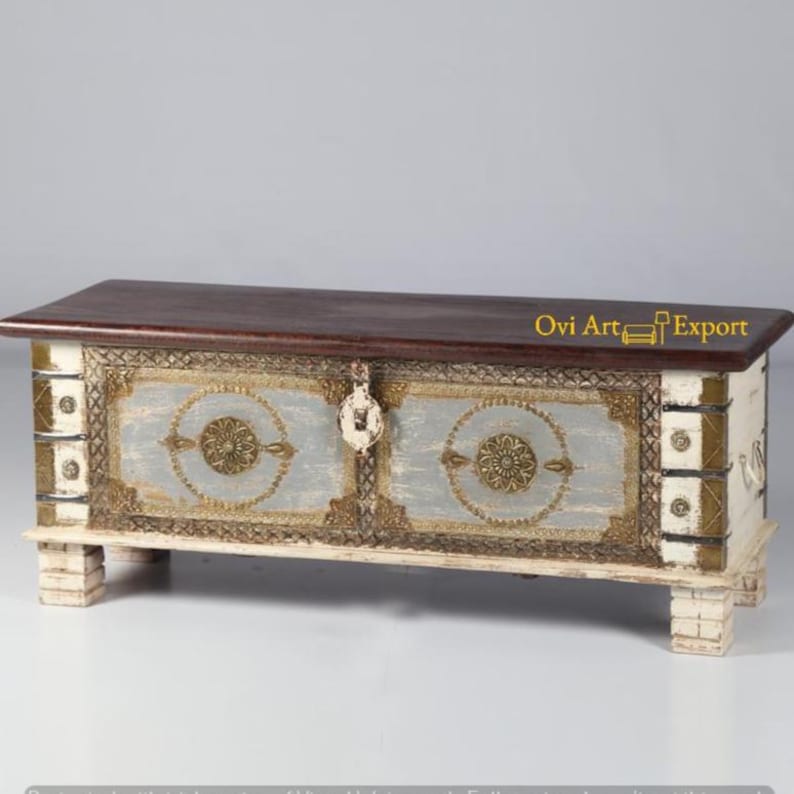 May include: A white wooden storage chest with gold accents and a dark wood top. The chest has two doors with ornate metal handles and a decorative design on the front. The text "Ovi Art Export" is visible on the top of the chest.