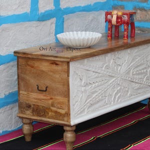 Peut inclure: Coffre de rangement en bois blanc avec des d&eacute;tails sculpt&eacute;s complexes sur le devant. Le coffre a un seul tiroir et repose sur quatre pieds tourn&eacute;s. Le coffre est sur un tapis ray&eacute; noir et rose. "Ovi Art and Export" est &eacute;crit sur le dessus du coffre.