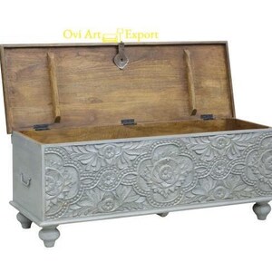 May include: A grey wooden storage chest with intricate carved floral designs. The chest has a hinged lid and sits on four turned legs.