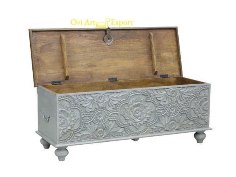 May include: A grey wooden storage chest with intricate carved floral designs. The chest has a hinged lid and sits on four turned legs.