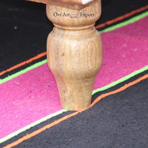 Peut inclure: Gros plan sur une patte de table en bois avec une finition naturelle. La patte repose sur un tapis ray&eacute; noir et rose. Le texte "Ovi Art Export" est visible sur le dessus de la table.