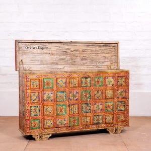 Puede incluir: Un cofre de madera con tapa con bisagras, pintado con un colorido patr&oacute;n de baldosas florales. El cofre es rectangular con un acabado blanco envejecido en la tapa y la base. El texto "Ovi Art Export..." es visible.
