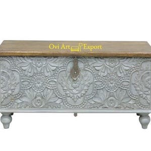 May include: A grey wooden storage chest with intricate carved floral designs and a wooden top. The chest has a metal latch and sits on four turned legs.