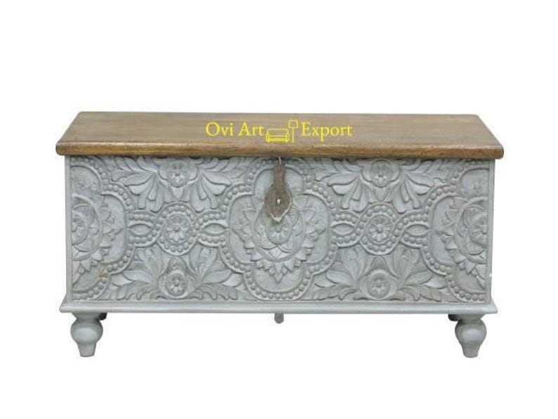 May include: A grey wooden storage chest with intricate carved floral designs and a wooden top. The chest has a metal latch and sits on four turned legs.