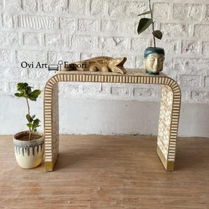 Può includere: Un tavolino da console bianco e dorato con un design curvo e intarsi intricati. Il tavolo è decorato con una piccola statuina di alligatore e una statua di testa di Buddha.