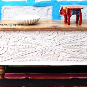 Peut inclure: Coffre de rangement en bois blanc avec des sculptures florales complexes. Le coffre a un dessus en bois naturel et quatre pieds tourn&eacute;s. Le coffre est pos&eacute; sur un tapis color&eacute; devant un mur bleu. Ovi Art Export est &eacute;crit sur le dessus du coffre.