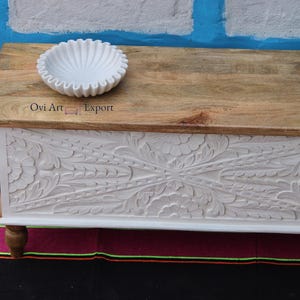 Peut inclure: Coffre de rangement en bois blanc avec des motifs floraux sculpt&eacute;s complexes. Le coffre a un dessus en bois naturel et repose sur quatre pieds. Un bol en c&eacute;ramique blanc est pos&eacute; sur le dessus du coffre.