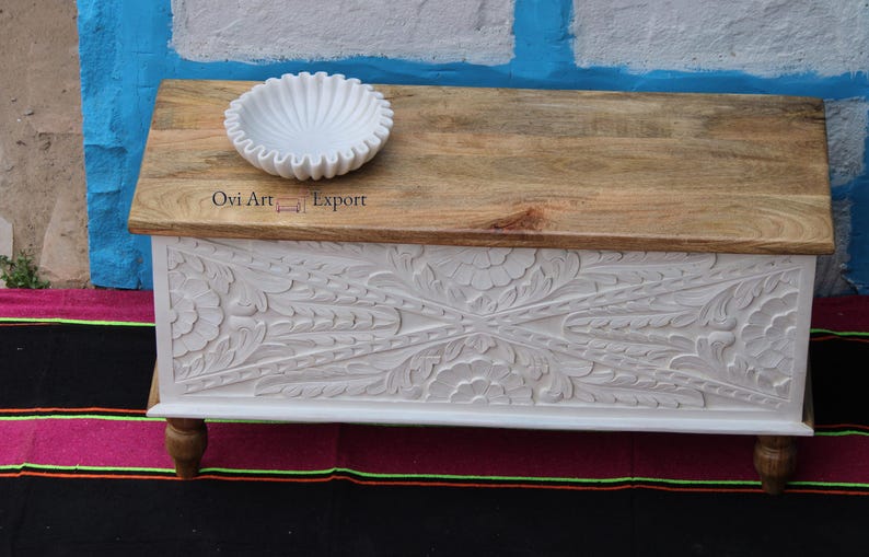 Peut inclure: Coffre de rangement en bois blanc avec des motifs floraux sculpt&eacute;s complexes. Le coffre a un dessus en bois naturel et repose sur quatre pieds. Un bol en c&eacute;ramique blanc est pos&eacute; sur le dessus du coffre.