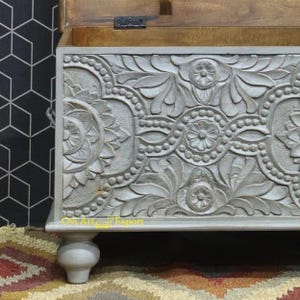 May include: A grey wooden storage chest with intricate carved floral designs. The chest has a hinged lid and sits on four turned legs.