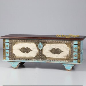 May include: A wooden storage chest with a distressed white and blue paint finish. The chest has ornate metal accents and two doors with brass handles. The chest is sitting on four small wheels.