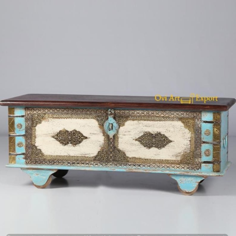 May include: A wooden storage chest with a distressed white and blue paint finish. The chest has ornate metal accents and two doors with brass handles. The chest is sitting on four small wheels.