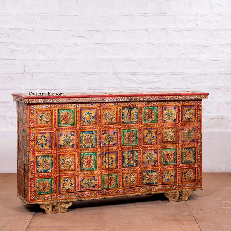 Puede incluir: Un mueble de madera pintado con colores vibrantes, con la parte superior y la base rojas. El mueble est&aacute; adornado con un patr&oacute;n repetitivo de cuadrados florales coloridos. El texto "Ovi Art Export" es visible. Descansa sobre patas cortas de color blanquecino.