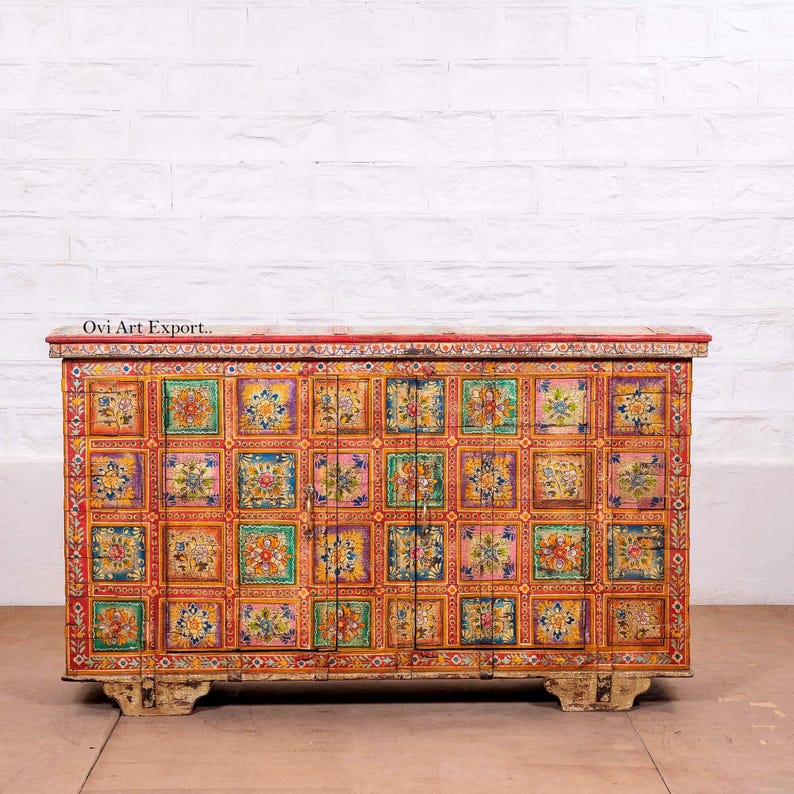 Puede incluir: Un mueble de madera pintado a mano con tapa y base rojas. El mueble presenta una cuadr&iacute;cula de coloridos dise&ntilde;os florales en cuadrados. El mueble tiene dos puertas y se apoya sobre patas cortas de color blanquecino. El texto "Ovi Art Export.." es visible.