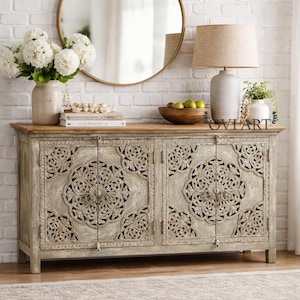 Puede incluir: Un aparador de madera gris desgastada con diseños florales tallados en las puertas. El aparador tiene una tapa de color marrón claro. Un espejo redondo dorado cuelga sobre el aparador, y una lámpara y un jarrón están encima. El aparador mide 150 cm de ancho.