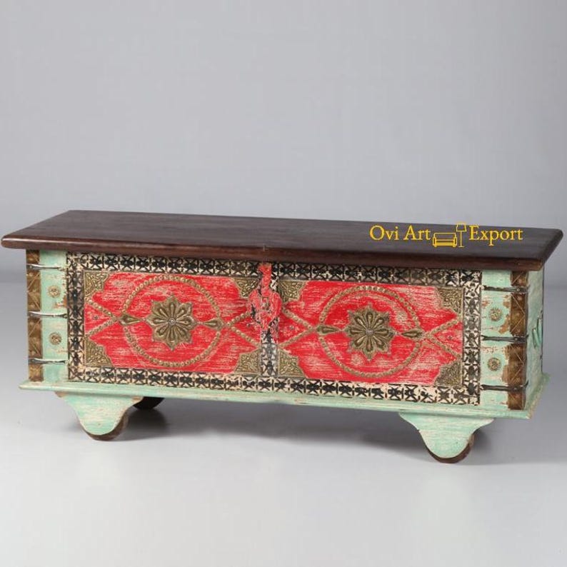 May include: A wooden storage chest with a distressed turquoise finish and intricate red and gold painted designs. The chest has a dark wood top and sits on four small wheels.