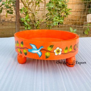 May include: Orange wooden tray with three legs and a painted design of a blue bird, green leaves, and a white flower. The tray is painted with a bright orange color and has a smooth, polished finish.