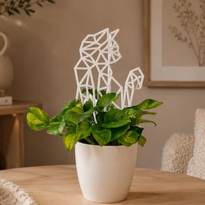 Könnte beinhalten: Ein weißer Keramiktopf enthält eine grüne Pflanze mit einem geometrischen weißen Katzen-Rankgitter. Die Pflanze steht auf einem Holztisch. Das Katzen-Rankgitter sorgt für eine dekorative Note.