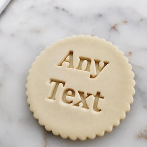 Op de afbeelding: Een ronde, lichtgekleurde koek met een geschulpte rand. De woorden "Any Text" zijn in het midden van de koek gedrukt. De koek ligt op een marmeren achtergrond.