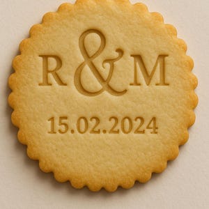 Könnte beinhalten: Ein runder, hellbrauner Keks mit gewelltem Rand. Der Keks ist mit den Buchstaben "R & M" und dem Datum "15.02.2024" geprägt. Der Keks ist ein Backwarenprodukt, wahrscheinlich für eine Hochzeit oder ein besonderes Ereignis.