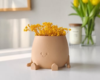 May include: A matte peach-colored ceramic planter shaped like a seated figure with a simple engraved face consisting of two circular eyes and a curved mouth, filled with a cluster of bright yellow dried flowers.