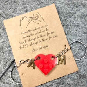 Op de afbeelding: Een rode hartvormige bedel met de letters "A" en "M" aan een ketting, bevestigd aan een kaart met de tekst "No matter where we go... One for you." De achtergrond is een grijze stof.