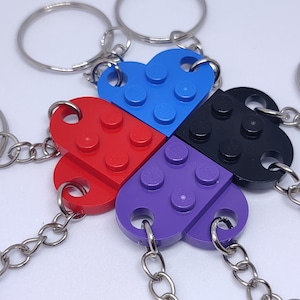 Op de afbeelding: Een close-up van vier hartvormige sleutelhangers in rood, blauw, zwart en paars. Elke sleutelhanger is gemaakt van in elkaar grijpende plastic blokken, met een metalen sleutelring en ketting. De sleutelhangers zijn in een bloemachtig patroon gerangschikt.