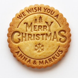 Può includere: Un biscotto natalizio rotondo e marrone dorato con bordo smerlato. Il biscotto è impresso con le parole "WE WISH YOU A MERRY CHRISTMAS" e i nomi "ANNA & MARKUS", insieme a un fiocco di neve e due piccoli alberi.
