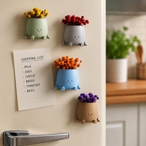 Op de afbeelding: Vier kleine, ronde, magnetische plantenbakjes in saliegroen, grijs, lichtblauw en beige, elk met een eenvoudig reliëfgezichtje en twee kleine voetjes. Ze bevatten gele, rode, oranje en paarse droogbloemen. Een boodschappenlijstje hangt ernaast.