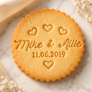 Peut inclure: Un biscuit rond à bords festonnés avec un motif en relief. Le texte "Mike & Allie" est centré, avec la date "11.06.2019" en dessous. Quatre petits cœurs sont gravés autour du texte, trois au-dessus et un en dessous.