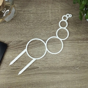 Op de afbeelding: Wit metalen draadcirkelontwerp met een reeks kleinere cirkels die met elkaar zijn verbonden om een ​​ketting te vormen. Het ontwerp heeft twee lange tanden die vanaf de onderkant uitsteken.