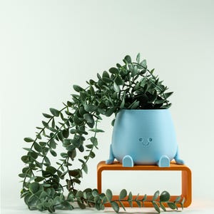 Op de afbeelding: Een lichtblauwe plantenpot met een glad, afgerond lichaam, kleine voetjes en een eenvoudig reliëfgezicht met twee ogen en een gebogen mond. Een hangende groene plant met kleine, ronde blaadjes hangt over de randen van de pot, die op een oranje rechthoekige standaard staat.