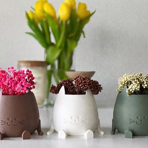 Op de afbeelding: Drie katvormige plantenbakken in bruin, wit en groen, elk met verschillende gedroogde bloemen. De plantenbakken hebben een minimalistisch ontwerp met een lachend gezicht en kleine pootjes. Gele tulpen staan in een glazen vaas op de achtergrond.