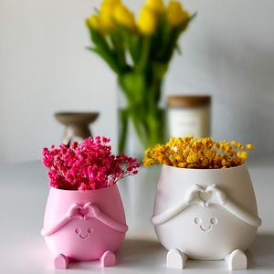 Op de afbeelding: Twee kleine, ronde plantenpotten met een matte afwerking, één roze en één wit. Elke pot heeft gebeeldhouwde armen die een hartvorm vormen boven een eenvoudig, gegraveerd lachend gezicht en twee kleine voetjes aan de basis. De roze pot bevat felroze droogbloemen, terwijl de witte pot gele droogbloemen bevat.