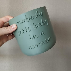 Puede incluir: Una maceta cilíndrica de color turquesa con la frase "nobody pots baby in a corner" grabada en relieve en el lateral. La maceta está sujeta por una mano y el fondo es de color neutro.