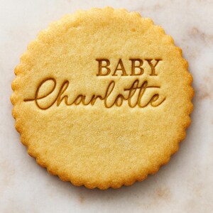 Könnte beinhalten: Ein runder, goldbrauner Keks mit gewelltem Rand. Der Keks ist mit den Worten "BABY Charlotte" in einer dekorativen Schriftart versehen, perfekt für eine Babyparty oder Geburtsanzeige.