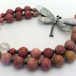 Könnte beinhalten: Eine zweireihige Halskette mit rosa und braunen Perlen, einem silbernen Libellenanhänger mit Strassstein und einem silbernen Schiebeverschluss.
