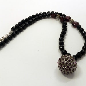 Könnte beinhalten: Eine schwarze Perlenkette mit einem silbernen Verschluss und einem großen, runden Anhänger, der mit violetten Strasssteinen besetzt ist.