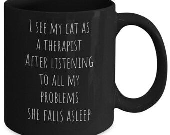 May include: Black coffee mug with white text that reads "I see my cat as a therapist after listening to all my problems she falls asleep."