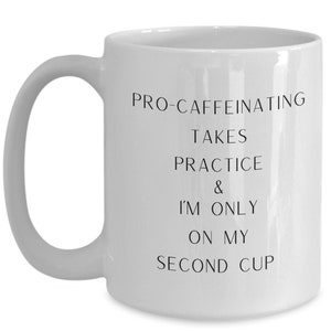 May include: White ceramic coffee mug with black text that reads "Pro-caffeinating takes practice & I'm only on my second cup."